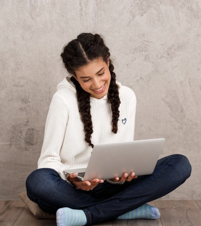 Beautiful african girl smiling, holding laptop, sitting on floor over beige background. Copy space.