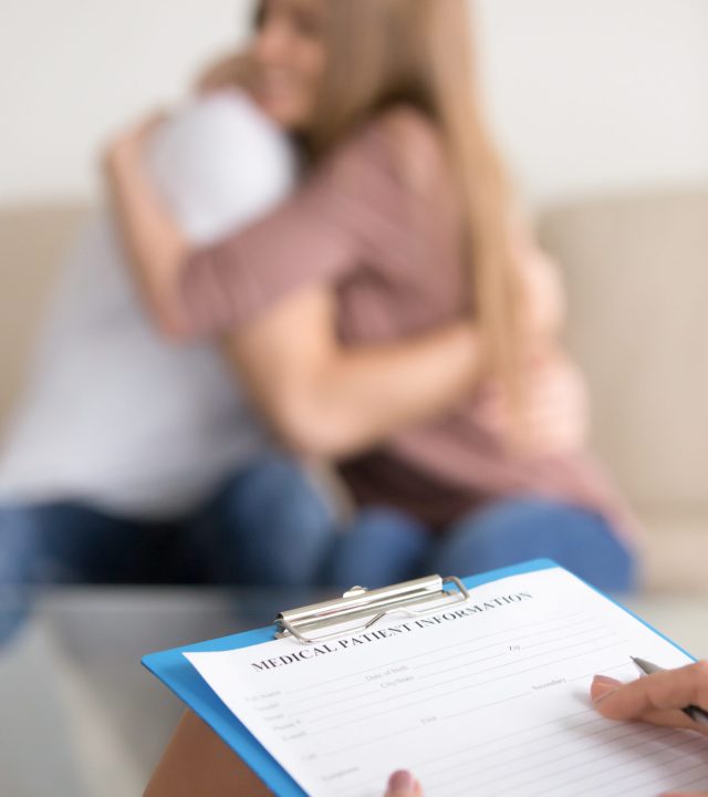 Close up of female psychotherapist holding clipboard, filling information about patients while couple embracing on couch in the background, doctor making notes in medical card in psychologists office